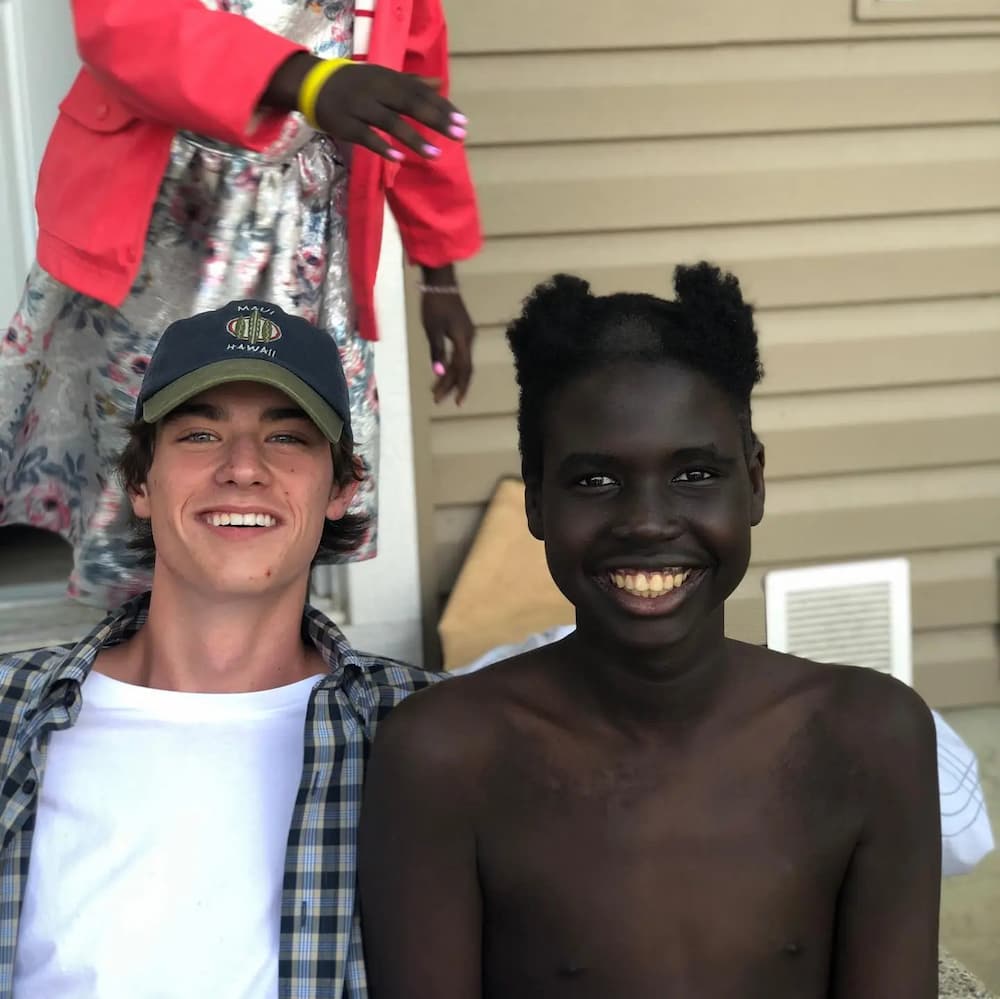 Two boys sit smiling, one with his head shaved to look like a clown
